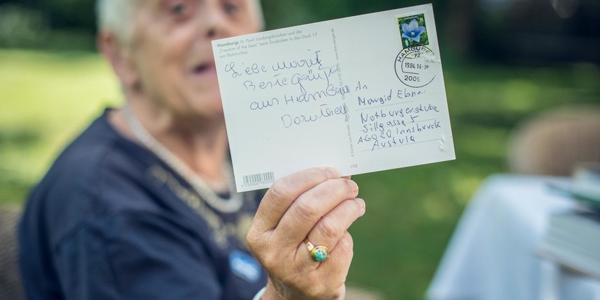 Eine ältere Frau hält eine Postkarte in die Kamera.