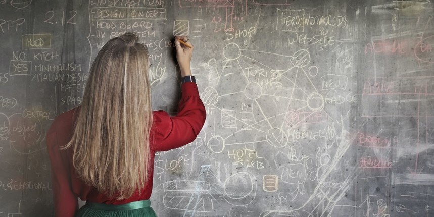 Eine blonde Frau schreibt mit Kreide an eine Tafel.